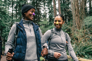 two women looking at each other and hiking in the forest with purple and yellow sunscreen on just their noses