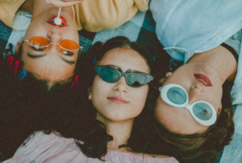 an aerial view of three women laying down on a checkered picnic blanket all wearing colorful sunglasses and smiling