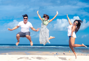 one man and two women that all wear sunglasses are jumping in the air excitedly with their arms up, they are on a beach with the ocean directly behind them 